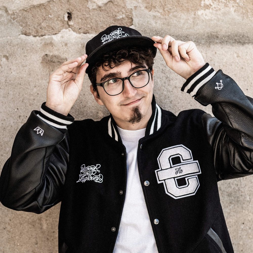 Auf dem Bild ist Flo zu sehen. Er trägt eine schwarze Cromatics-Jacke und greift sich an sein Cap