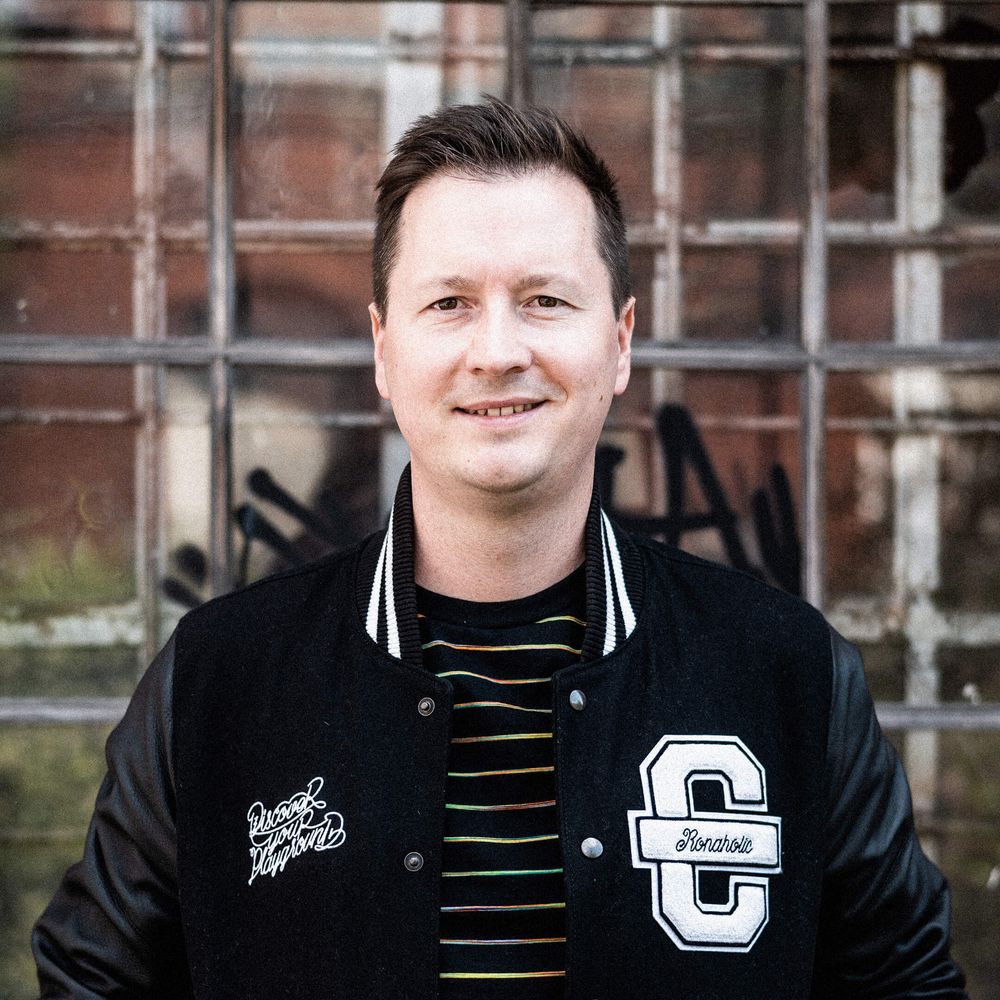 # Auf dem Bild ist Ron zu sehen. Er trägt eine schwarze Cromatics-Jacke und lächelt verschmitzt.