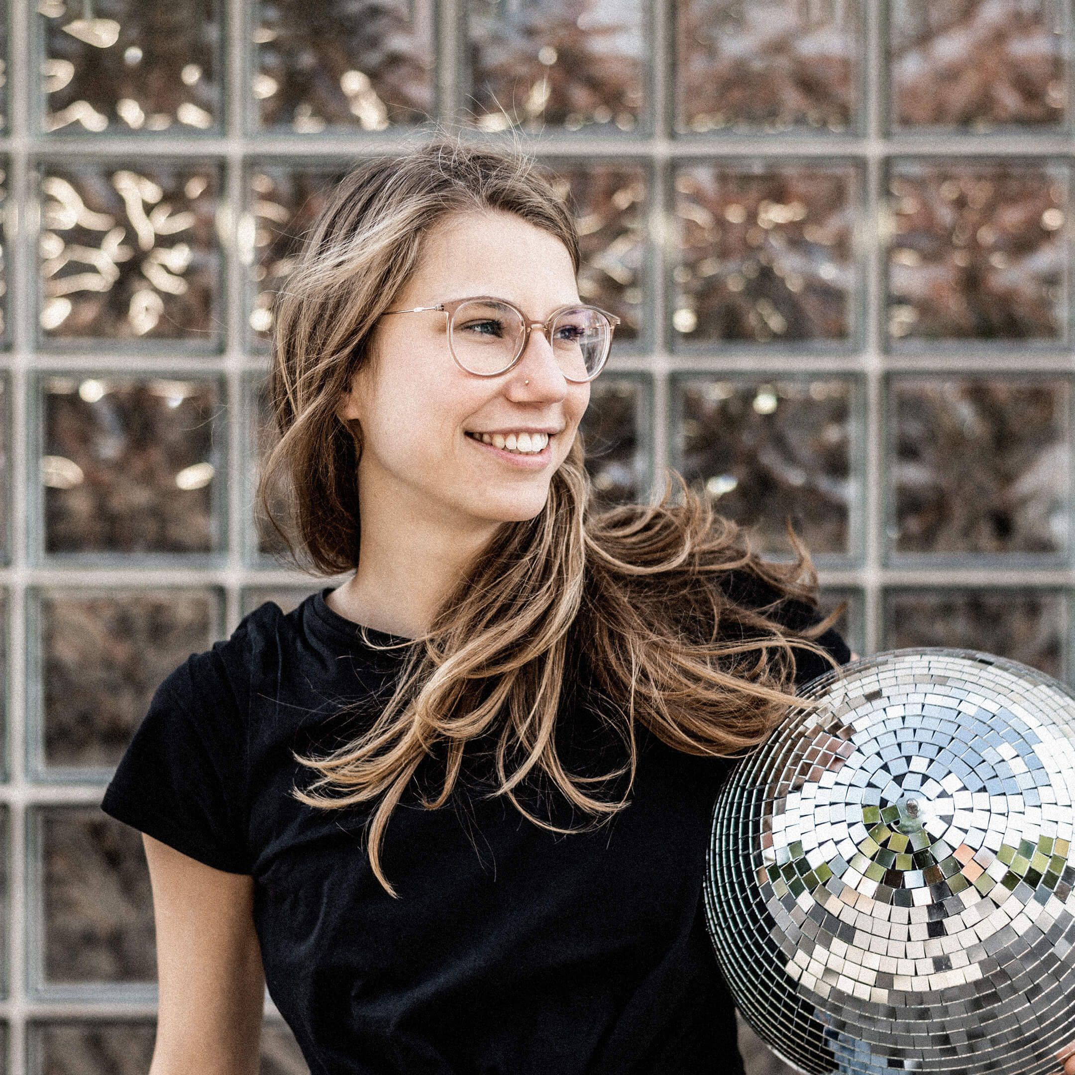 Auf dem Bild ist Ella zu sehen. Sie trägt eine Diskokugel unter dem Arm und schaut fröhlich.