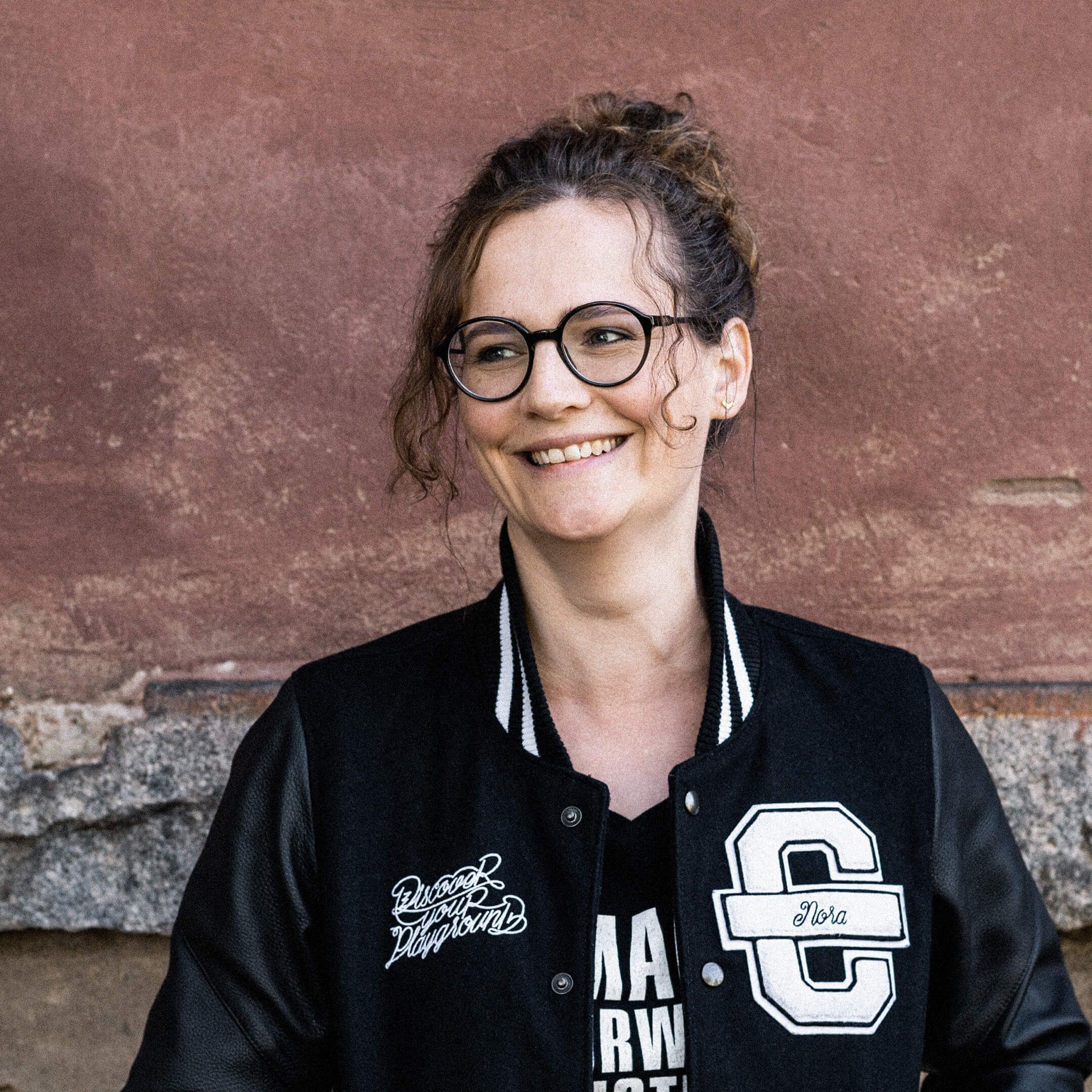 Hier ist Nora zu sehen. Sie schaut lächelnd nach links und trägt eine schwarze Cromatics-Jacke.